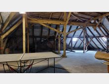 Ein geräumiger Dachboden mit sichtbaren Holzbalken und einer Holzbalkenkonstruktion, teils bedeckt mit einer Schutzverkleidung. Im Vordergrund steht ein Tischtennistisch, während durch kleine Fenster an den Seiten Licht einfällt.