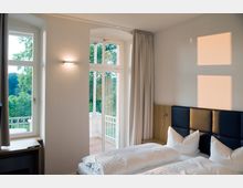 Ein hell eingerichtetes Schlafzimmer mit einem Doppelbett, einem gepolsterten Kopfteil und weißen Bettdecken. Große Fenster und eine verglaste Balkontür lassen Tageslicht herein und bieten einen Blick auf einen grünen Außenbereich.