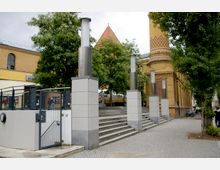 Außenansicht eines historischen gelben Backsteingebäudes mit Ornamenten, flankiert von Bäumen. Im Vordergrund führen Steinstufen mit modernen Pfeilern aus Metall und Beton nach oben, daneben ein Gittertor und ein gepflasterter Gehweg.