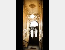 Glasverzierte Eingangstür mit floralem Muster, die zu einem Treppenhaus führt, dessen Wände mit hellen Stuckverzierungen und goldenen Akzenten geschmückt sind. Im Hintergrund ist eine geöffnete Holztür mit Glaseinsätzen zu sehen.