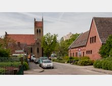 Straßenszene in einem Wohngebiet mit gepflasterter Fahrbahn, geparkten Autos und umliegenden Gebäuden aus rotem Backstein. Im Hintergrund ist eine Kirche mit zwei Türmen und roten Ziegeln zu sehen, umgeben von Bäumen.
