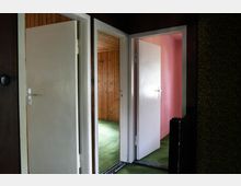 Ein Flur mit geöffneter Tür, die zu einem Raum mit holzvertäfelten Wänden führt, sowie eine weitere Tür, die einen Raum mit rosa gestrichenen Wänden zeigt. Der Boden ist mit grünem Teppich ausgelegt, und an den Wänden ist teils gemusterte Tapete sichtbar.