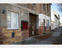Straßenansicht eines alten, teils heruntergekommenen Gebäudes mit grauer Fassade, gelb-roter Klinkerverkleidung und Graffiti an den Wänden. Ein großes Holztor, eine Fensterscheibe mit Jalousien und ein leeres Plakatrahmen sind sichtbar; der Gehweg ist mit Herbstlaub bedeckt.