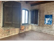 Raum mit einer Wand aus rohen Ziegelsteinen, drei Fenstern (eines geöffnet mit Holzläden), einem Holzstuhl und einem abstrakten, blau-weißen Kunstwerk an der Wand. Die Decke besteht aus Holzbalken, und der Boden ist schlicht gestaltet.