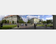 Panoramaaufnahme eines belebten Stadtplatzes mit historischen und modernen Gebäuden. Links ein großes Gebäude mit rotem Dach und klassischer Fassade, rechts eine gelb-blaue Büro- oder Geschäftshausfassade, umgeben von Grünflächen, Bäumen und Spaziergängern.