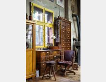 Ein rustikal eingerichteter Raum mit Holzfußboden, alten Holzschränken mit zahlreichen kleinen Schubladen, einem gelben Fensterrahmen, einem hölzernen Hocker und einem braunen Ledersessel. Im Hintergrund sind eine Glasvitrine, eine Werkstattlampe und größere Industrieelemente sichtbar.