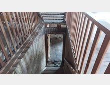 Holztreppe mit Geländer führt in einen Innenraum mit Ziegelmauern und Ziegelfußboden, umgeben von Holz- und Backstein-Elementen in einer rustikalen Umgebung.