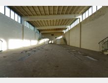 Innenansicht einer verlassenen Industriehalle mit hohen Decken, rechteckigen Fenstern entlang der oberen Wände und einem Boden mit eingelassenen Gruben. An beiden Seiten führen Treppen mit Metallgeländern zu erhöhten Plattformen.