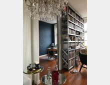 Innenraum einer Wohnung mit Holzfußboden, einem modernen Bücherregal, das eine Wand einnimmt, und einem hellen Arbeitsbereich im Hintergrund mit einem Schreibtisch und einem blauen Akzent an der Wand. Im Vordergrund ein Kronleuchter und zwei kleine Beistelltische mit Deko-Elementen.