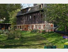 Ein zweistöckiges Wohnhaus mit dunkler Fassade und gemauertem Erdgeschoss inmitten eines Gartens mit Rasen, blühenden Bäumen und Blumenbeeten. Im Vordergrund stehen gestreute Gartenmöbel sowie ein Kanu und ein kleiner Anhänger.