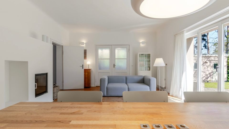 Helles, modern eingerichtetes Wohnzimmer mit Holztisch im Vordergrund, grauem Sofa, eingebautem Kamin und weißer Deckenleuchte. Große Fensterfront mit Vorhängen und Terrassentür lässt Tageslicht herein und bietet Blick ins Grüne.