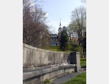 Historisches Backsteingebäude mit Turm und dekorativer Spitze, umgeben von Bäumen in einem Park mit leicht geneigtem Hügel und einer steinernen Mauer im Vordergrund.