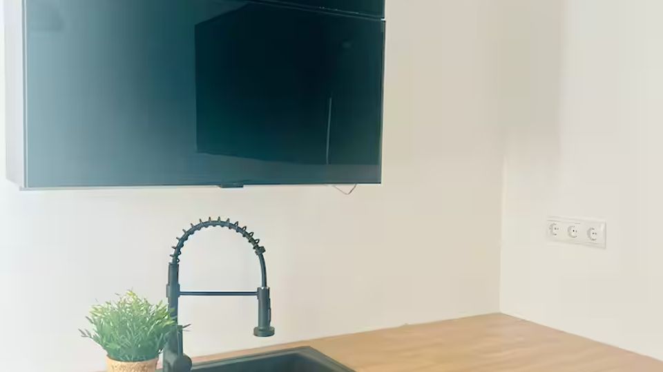 Moderne Küchenzeile mit Holzarbeitsplatte, schwarzer Spüle und hohem schwarzen Federhahn, dazu ein kleiner Pflanztopf auf der Abtropffläche. Darüber hängen zwei große schwarze, hochglänzende Hängeschränke an einer weißen Wand.