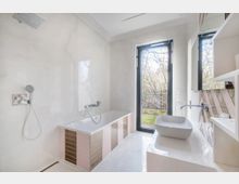 Helles Badezimmer mit freistehender Badewanne, rechteckigem Waschbecken und großer Glastür, die ins Freie führt. Die Wände und der Boden sind in weißen und beigefarbenen Tönen gehalten, mit dekorativen Fliesen und Blick auf einen Gartenbereich.