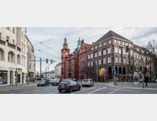Straßenkreuzung mit einem historischen roten Backsteingebäude mit Turm und Uhr rechts im Bild sowie einem weiteren großen Gebäude daneben. Im Vordergrund fließender Autoverkehr, Straßenlaternen und Fußgänger auf Gehwegen, während der Himmel bedeckt ist.