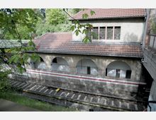 Ein altertümliches Gebäude mit roten Dachziegeln und Bogenarkaden im Erdgeschoss, hinter denen vergitterte Fenster sichtbar sind. Im Vordergrund verlaufen Bahngleise, umgeben von Bäumen und dichter Vegetation.