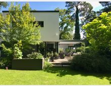 Moderne Einfamilienhaus mit weißer Fassade, großen Glasfenstern und einer überdachten Holzterrasse, umgeben von einem gepflegten Garten mit Rasen, Bäumen und Sträuchern. Im Hintergrund sind hohe Bäume zu sehen, die eine grüne und ruhige Umgebung schaffen.