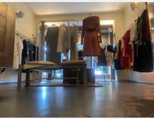 Ein kleiner, heller Bekleidungsladen mit einer Schaufensterpuppe im Vordergrund, die ein gemustertes Oberteil und einen braunen Rock trägt. Auf beiden Seiten des Raums hängen Kleidungsstücke auf Kleiderstangen, während Tageslicht durch die Fenster im Hintergrund fällt.