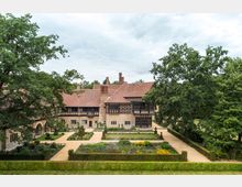 Historische Hofanlage mit Fachwerkelementen und roten Ziegeldächern, umgeben von üppigem Grün. Im Vordergrund befindet sich ein gepflasterter Innenhof mit symmetrischen Blumenbeeten und geschnittenen Hecken.