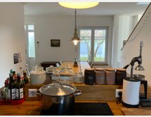 Moderne Küche mit einer hölzernen Arbeitsplatte, auf der Gläser mit Vorräten, Flaschen und Gewürzdosen stehen. Im Hintergrund ein Esstisch mit Stühlen vor einem Fenster, das auf einen Garten mit Bäumen blickt; daneben eine Treppe, die nach oben führt.