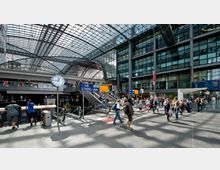 Eine moderne Bahnhofshalle mit Glasdach und großen Fensterfronten. Menschen bewegen sich zwischen Etagen über Rolltreppen und Wegebrücken, während eine große analoge Uhr und Beschilderungen für Bahnsteige und Services sichtbar sind.