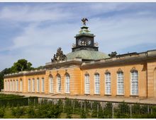 Historisches Gebäude mit gelb-orangefarbener Fassade, großen weiß gerahmten Fenstern und einem grünen Kuppeldach mit einem Glockenturm, der von einer Adlerstatue gekrönt ist. Im Vordergrund ist eine Gartenanlage mit Hecken und Bäumen zu sehen.