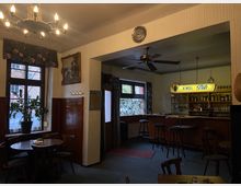 Gemütliches Interieur einer traditionellen Kneipe mit Holzmöbeln, einem runden Tisch und Stühlen im Vordergrund. Im Hintergrund eine Bar mit Holztheke, Barhockern und einem beleuchteten "Kindl-Pils"-Schild, sowie dekorativen Elementen wie einem Kronleuchter und einem Ventilator an der Decke.