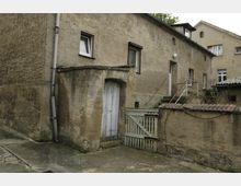Ein Hof mit alten, verputzten Gebäuden aus hellbraunem Beton. Sichtbar sind eine kleine Holztür in einem Nebenbau, ein verwittertes Holztor und einfache, teils verglaste Fenster mit dunklen Fensterrahmen.