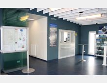 Innenraum einer modernen Touristeninformation mit weiß-blauen Wänden und blauem Bodenbelag. Zu sehen sind ein Informationsschalter mit Glasabtrennung, ein Stehtisch, ein Schaukasten mit Broschüren sowie ein beleuchtetes Deckenpanel.