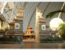 Innenansicht einer großen Halle mit gewölbter, gitterartiger Decke und einem balinesischen Tor aus Stein in traditionellem Design. Dahinter steht ein mehrstöckiges Gebäude mit Strohdach, umgeben von Pflanzen und dekorativen Elementen.