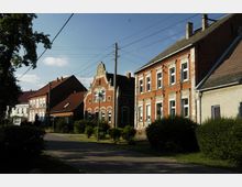 Straßenansicht in einer Wohngegend mit historischen Backsteinhäusern, verzierten Fassaden und roten Ziegeldächern, umgeben von kleinen Vorgärten und Büschen bei Tageslicht. Ein Gehweg führt entlang der Häuser, und Stromleitungen verlaufen über die Straße.