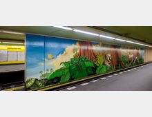 U-Bahn-Station mit dem Namen „Bismarckstraße“, gelb-grünes Farbschema, Wand mit farbenfrohem Wandgemälde, das eine Dschungelszene mit großen Blättern, einem Baumstamm, einer Cartoon-Figur und anderen Details zeigt. Der Bahnsteig ist grau mit wartenden Gleisen.