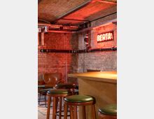 Ein Barbereich mit Hockern mit grünen Polstersitzen an einer Holztheke, einer Backsteinwand im Hintergrund und einem roten Neonlicht mit der Aufschrift „BIER TAP“. Sichtbare Rohrleitungen an der Decke und ein Ledersessel im hinteren Bereich vervollständigen das industrielle Design.