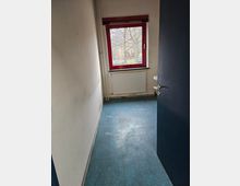 Schmaler Raum mit blauem Bodenbelag, weißen Wänden und einem roten Fensterrahmen. Unter dem Fenster befindet sich ein Heizkörper, und rechts ist ein kleiner Teil eines Waschbeckens sichtbar.