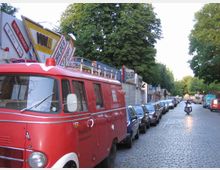 Eine gepflasterte Straße mit parkenden Autos auf beiden Seiten, vorn links ein rotes Vintage-Fahrzeug mit Dachleiter und rechts ein Motorradfahrer. Im Hintergrund sind mehrere Bäume, Geschäfte mit Werbeschildern und ein Gebäude sichtbar.