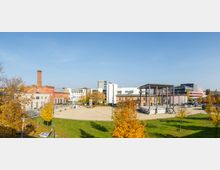 Weitläufiger Platz mit einem gemischten Ensemble aus historischen Backsteingebäuden und modernen Bauten, darunter ein Schornstein und eine offene Stahlkonstruktion. Im Vordergrund befinden sich Grünflächen und Bäume mit Herbstlaub, während im Hintergrund moderne Bürogebäude sichtbar sind.