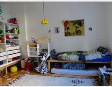 Ein Kinderzimmer mit hellem Holzboden, einem einfachen Holzbett mit bunter Bettwäsche, kleinen Regalen mit Spielsachen und Büchern sowie einem weißen Teppich mit geometrischem Muster. An der Wand hängen Bilder, und ein großer Plüschhund sitzt neben dem Bett.