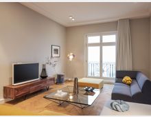 Wohnzimmer mit moderner Einrichtung, darunter ein graues Sofa, ein gelbes Sitzmöbel und ein Couchtisch mit Glasplatte. Ein niedriger Holzschrank mit einem Fernseher steht an der Wand, daneben eine dekorative Lampe und ein Balkon mit Glas- und Metalltür im Hintergrund.