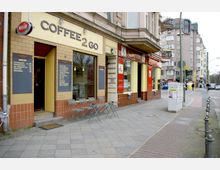 Straßenszene mit einem kleinen Café namens "Coffee 2 Go" auf der linken Seite, mit gelber Fassade, schwarzen Tafeln und kleinen Sitzgelegenheiten. Daneben befindet sich ein Geschäft mit einer rot-gelben Fassade, während im Hintergrund städtische Wohngebäude und eine Straße mit Fußgängern und Autos sichtbar sind.
