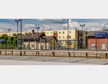 Bahnhofsumgebung in Lübben (Spreewald), mit einem Bahnsteig, einem niedrigen Gebäude mit Ziegeldach, modernen Wohnhäusern im Hintergrund und einem blauen Stationsschild. Strommasten und Lampen prägen die Szenerie, die durch Zäune abgegrenzt ist.