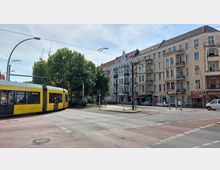 Straßenkreuzung in einer städtischen Umgebung mit einer gelben Straßenbahn im Vordergrund, die um eine Kurve fährt. Im Hintergrund befinden sich mehrstöckige Altbauten mit Balkonen und Geschäften im Erdgeschoss, flankiert von Bäumen und Gehwegen.