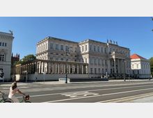 Historisches Gebäude mit klassizistischer Architektur, grauer Fassade und Säuleneingang, flankiert von weiteren historischen Gebäuden. Im Vordergrund eine Straße mit Linienmarkierungen und einer Person auf einem Fahrrad; blauer Himmel im Hintergrund.