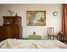 Schlafzimmer mit Retro-Einrichtung: Ein Holzbuffet mit Glasvitrine steht links, darauf Dekoobjekte. An der Wand hängen ein gerahmtes Landschaftsgemälde und eine Uhr. Rechts befinden sich ein Stuhl und ein kleiner Tisch mit einem dekorativen Gefäß. Im Vordergrund ist ein Bett mit heller Decke sichtbar.