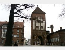 Backsteingotisches Stadttor mit spitzem Dach und weißen Fensterverzierungen aus dem Mittelalter, flankiert von angrenzenden Ziegelgebäuden, auf einer gepflasterten Straße mit kahlen Bäumen im Vordergrund. Im Hintergrund sind weitere Gebäude und eine belebte Straße zu sehen.