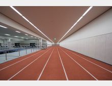 Ein Indoor-Leichtathletikbereich mit einer roten Laufbahn, die von weißen Linien unterteilt ist, und einer hellen Decke mit eingebauten Lichtpanelen. Rechts verläuft eine einfache Wand, während links ein abgetrennter Bereich mit Stuhlreihen und Bühnenkonstruktionen sichtbar ist.