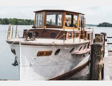 Ein historisches Holzboot mit weißem Rumpf und einem Aufbau aus dunklem Holz liegt an einem hölzernen Steg festgemacht. Im Hintergrund ist ein See mit bewaldetem Ufer und trübem Himmel zu sehen.
