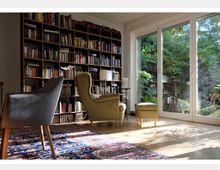 Wohnzimmer mit großer Bücherwand auf der linken Seite, zwei gemütlichen Sesseln (einer in Grau, einer in Grün mit passendem Hocker) und einem Teppich mit orientalischem Muster. Rechts befinden sich bodentiefe Fenster, die viel Licht hereinlassen und einen Blick auf einen grünen Garten bieten.