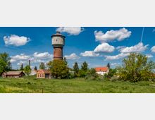 Eine ländliche Szene mit einem historischen Wasserturm aus Backstein und Beton in der Mitte, umgeben von grünen Wiesen, Bäumen und mehreren kleineren Gebäuden mit roten Ziegeldächern. Der Himmel ist blau mit einigen weißen Wolken.
