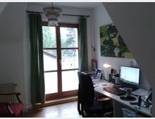 Arbeitszimmer mit Schreibtisch, Computer und Büromaterial. Im Hintergrund ein großes Fenster mit grünem Vorhang und Blick auf eine Terrasse mit Bäumen im Freien. Links steht ein gepolsterter Stuhl, und rechts hängt ein Bild mit Blumenmotiv an der Wand.