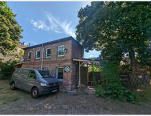 Backsteingebäude auf einem Grundstück mit Kopfsteinpflaster, umgeben von mehreren großen Bäumen. Vor dem Gebäude steht ein grauer Transporter, und an der rechten Seite des Hauses ist ein überdachter Bereich mit Rankenpflanzen erkennbar.